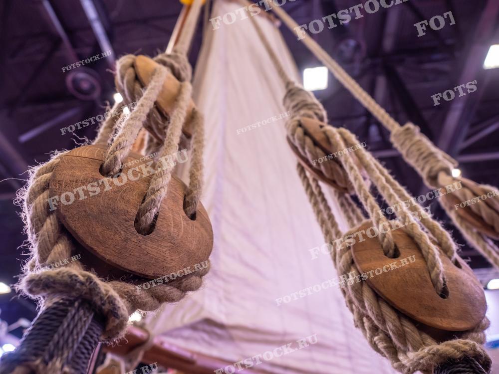 На фотографии крупным планом изображены такелаж и паруса на старинном парусном корабле. Видны деревянные блоки и веревки, используемые для управления парусами, иллюстрирующие морское наследие и традиции кораблестроения.